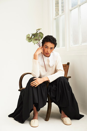 A woman with short curly hair sits on a wooden chair in a white sweater, black skirt, and beige flats, holding green leaves.
