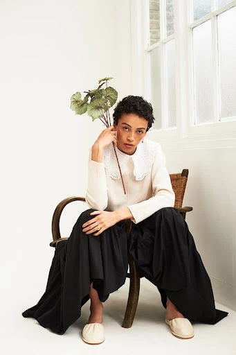 A woman with short curly hair sits on a wooden chair in a white sweater, black skirt, and beige flats, holding green leaves.