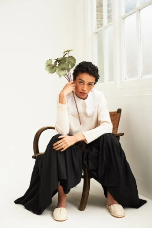 A woman with short curly hair sits on a wooden chair in a white sweater, black skirt, and beige flats, holding green leaves.