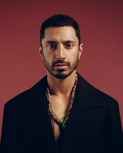Portrait of Riz Ahmed in a black coat against a maroon backdrop, showcasing his stylish necklace and serious expression.