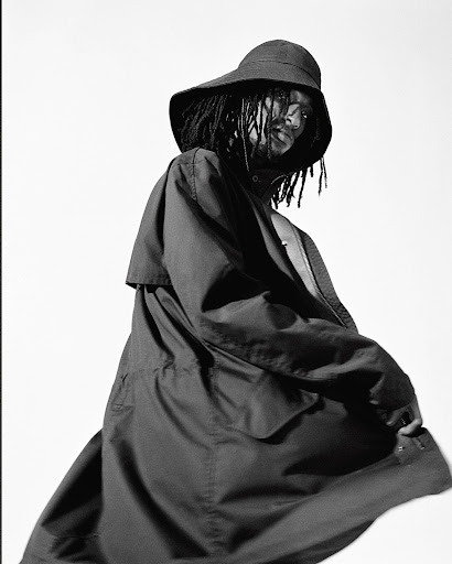Black and white portrait of a person with dreadlocks wearing a wide-brimmed hat and a dark coat against a light, neutral background.