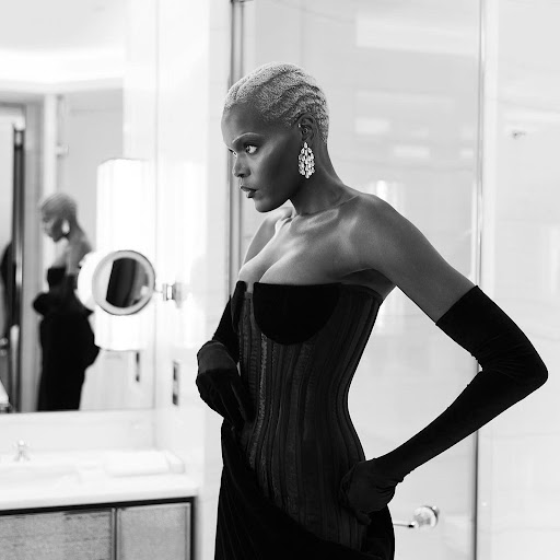 A woman with short white hair poses in a black form-fitting dress and gloves, glancing away from the camera inside a bathroom.