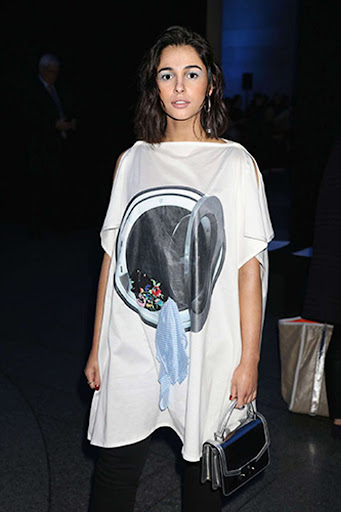 Woman wearing a white dress with a washing machine graphic, black pants, and a purse, standing indoors with a dark background.