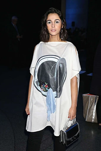 Woman wearing a white dress with a washing machine graphic, black pants, and a purse, standing indoors with a dark background.