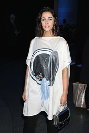 Woman wearing a white dress with a washing machine graphic, black pants, and a purse, standing indoors with a dark background.