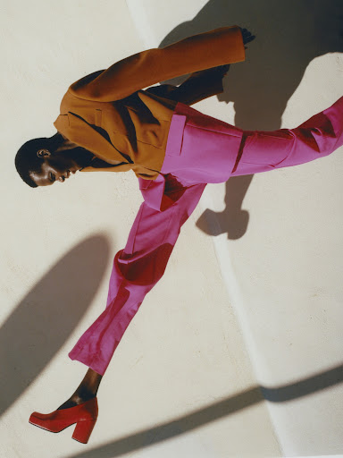 Stylish woman in a brown blazer and pink pants strikes a dynamic pose on a white surface, complemented by red heels and a strong shadow.