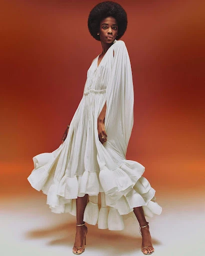 A Black female fashion model poses in a flowing, white gown with cape-like sleeves and strappy heels against a warm orange background.