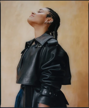 Profile portrait of a woman in a black leather jacket with braided hair and ear cuffs against a warm textured wall.