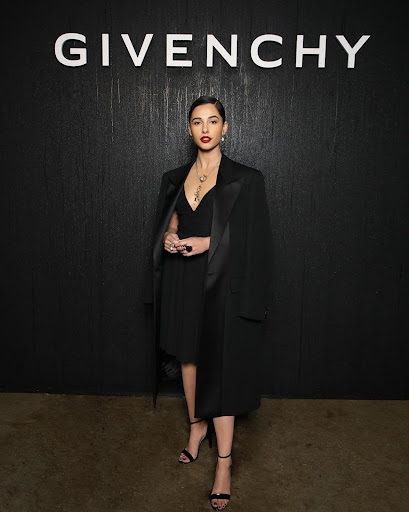 Naomi Scott at a fashion event, posing in a black dress and coat in front of a Givenchy backdrop, showcasing elegant style and fashion.