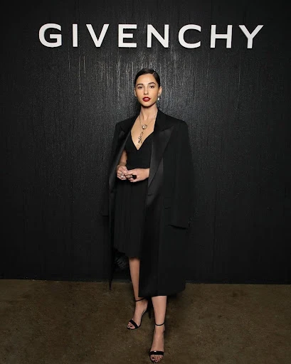 Naomi Scott at a fashion event, posing in a black dress and coat in front of a Givenchy backdrop, showcasing elegant style and fashion.
