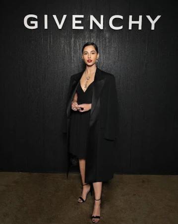 Naomi Scott at a fashion event, posing in a black dress and coat in front of a Givenchy backdrop, showcasing elegant style and fashion.
