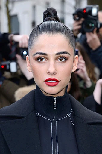 Close-up portrait of a beautiful adult woman with red lipstick, dark hair in a bun, and wearing a black coat; photographers in background.