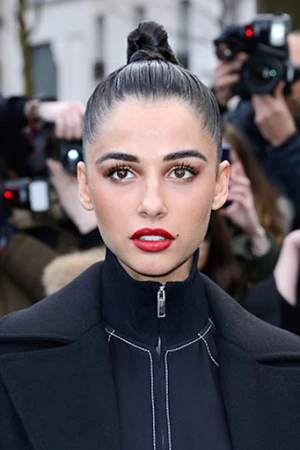 Close-up portrait of a beautiful adult woman with red lipstick, dark hair in a bun, and wearing a black coat; photographers in background.