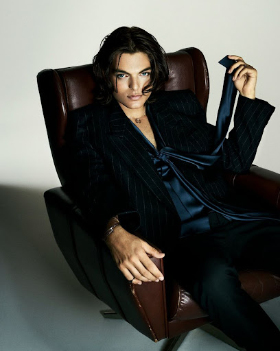 Portrait of a young adult male with long hair sitting in a leather chair wearing a suit jacket and silk shirt in a studio setting.