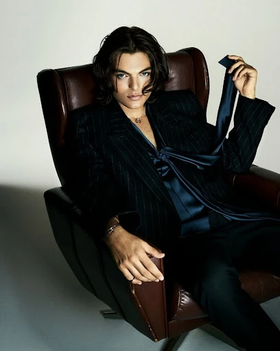 Portrait of a young adult male with long hair sitting in a leather chair wearing a suit jacket and silk shirt in a studio setting.