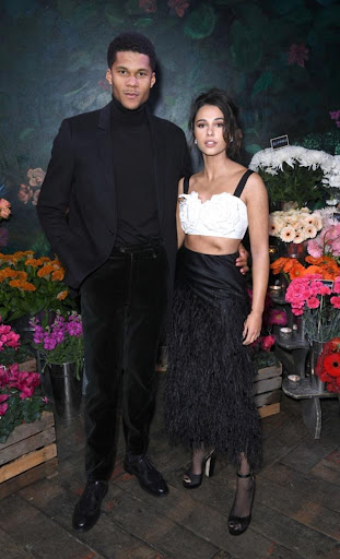 A couple poses in front of flowers: a man in a black suit and turtleneck, and a woman in a white top and black feathered skirt.