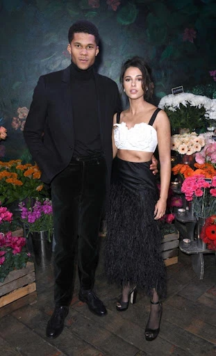 A couple poses in front of flowers: a man in a black suit and turtleneck, and a woman in a white top and black feathered skirt.