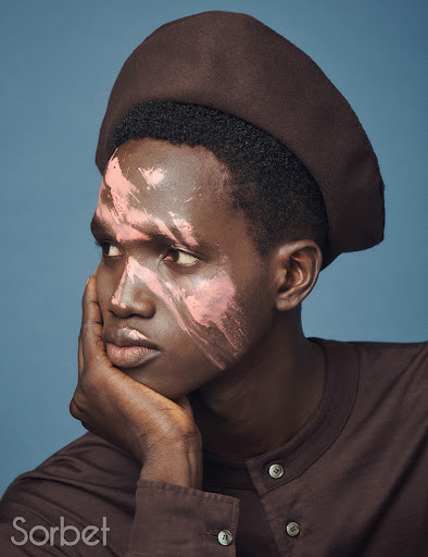 Portrait of a Black man with light pink face makeup, wearing a brown beret and collared shirt, set against a blue background.