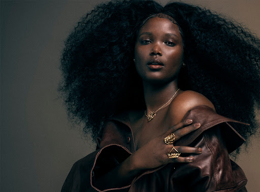 Portrait of a beautiful dark-skinned woman with natural hair wearing gold jewelry and a brown leather jacket in a neutral studio setting.