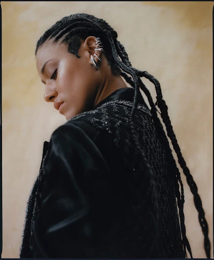 Close-up of a woman in a black jacket with braided hair, looking down with fair skin and silver hoop earrings against a beige background.