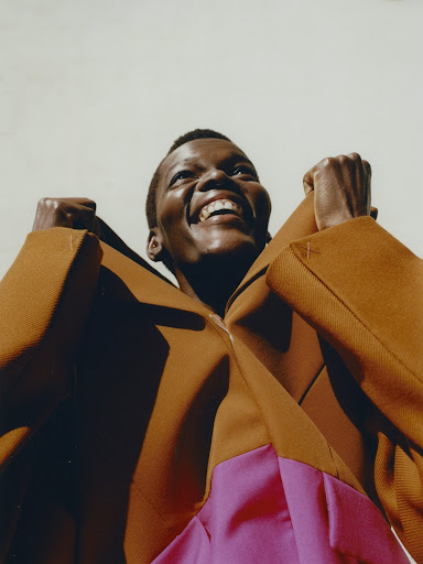 Low angle portrait of a Black woman in a brown and pink color block outfit, smiling with fists up, against a pale background.