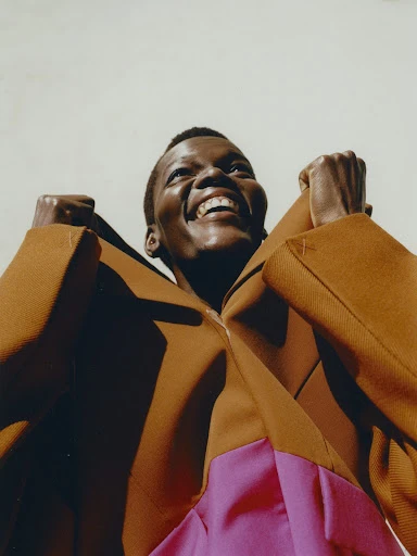 Low angle portrait of a Black woman in a brown and pink color block outfit, smiling with fists up, against a pale background.