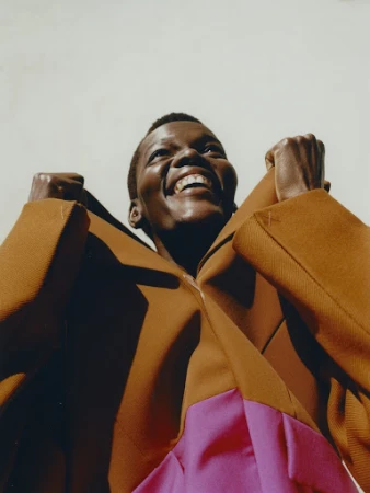 Low angle portrait of a Black woman in a brown and pink color block outfit, smiling with fists up, against a pale background.