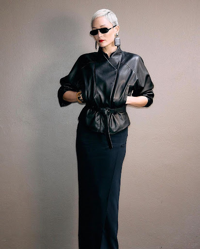 Adult female fashion model with short platinum hair poses in a black leather jacket and long black skirt against a neutral backdrop.