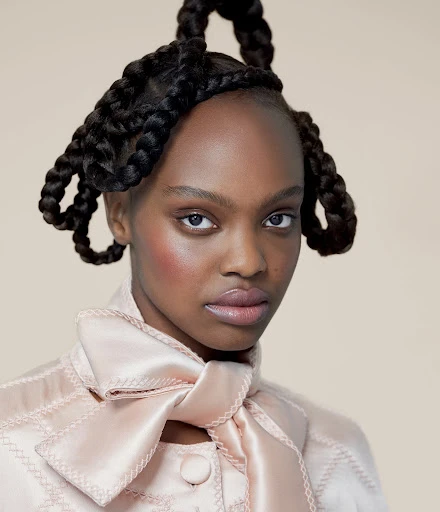 Close-up portrait of a Black woman with elaborate braided updo hairstyle and a light pink jacket with a large bow, against a neutral backdrop.