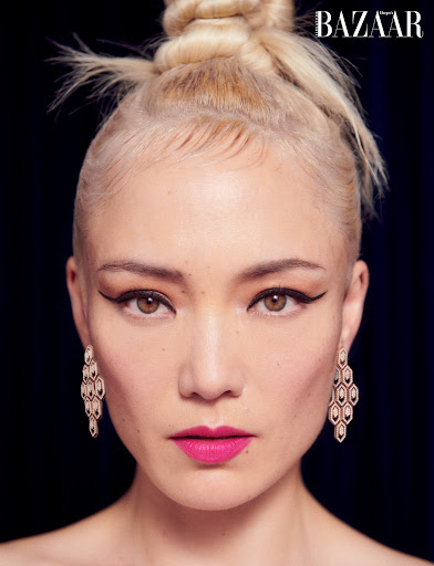 Close-up portrait of a woman with blonde hair in a high bun, winged eyeliner, and geometric diamond earrings against a dark background.