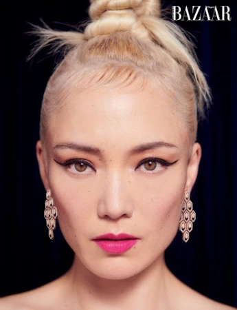 Close-up portrait of a woman with blonde hair in a high bun, winged eyeliner, and geometric diamond earrings against a dark background.