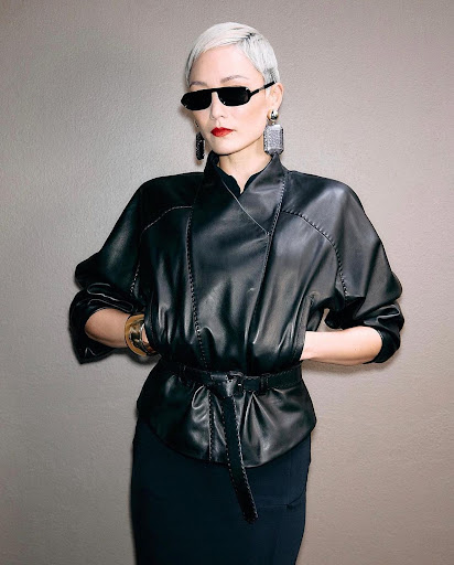 Short-haired model in black leather jacket and sunglasses against a solid grey wall