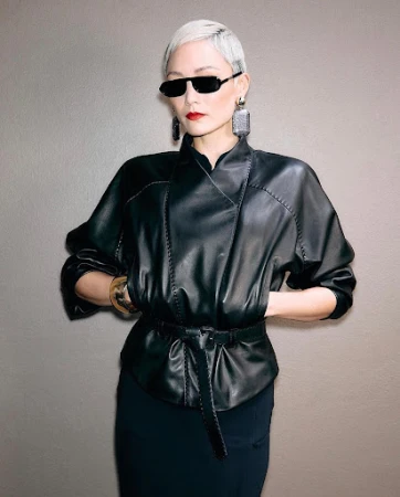 Short-haired model in black leather jacket and sunglasses against a solid grey wall