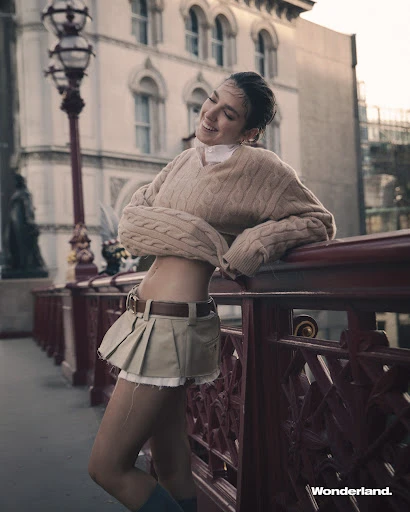 Smiling adult woman in a sweater and skirt poses outdoors in an urban environment with a bridge and buildings in the background.