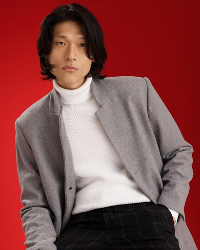 Man with dark hair wearing a grey coat and white turtleneck sweater against a solid red background