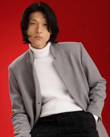 Man with dark hair wearing a grey coat and white turtleneck sweater against a solid red background