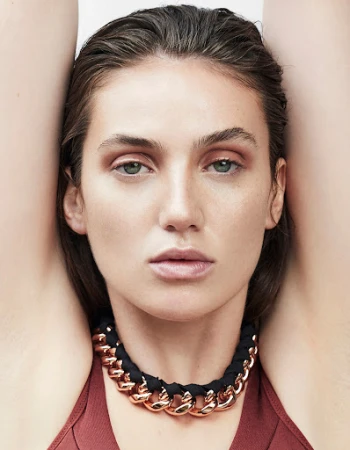 Close-up portrait of a woman with wet hair wearing a chunky rose gold chain necklace and a burgundy top against a plain light background