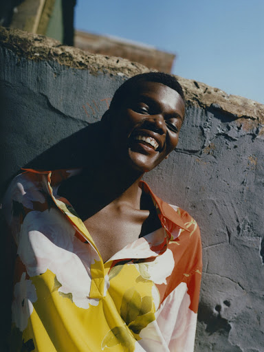 Smiling Black woman wearing a vibrant floral print shirt leaning against a grey textured outdoor wall under bright sunlight.