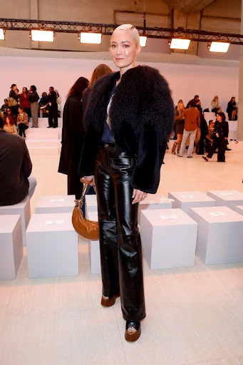 Fashionable woman with short bleached hair poses in black coat and shiny pants at indoor event with white cube seating, medium shot.