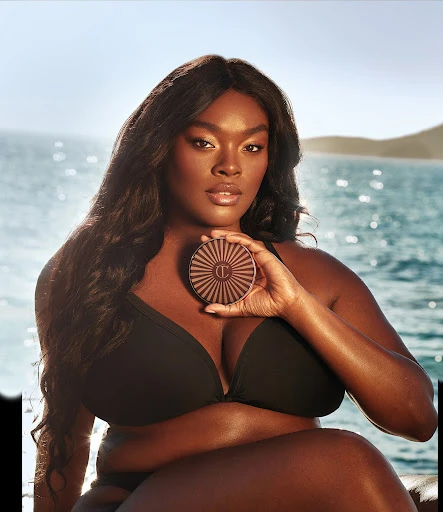 Woman in black bikini holds bronzer by ocean with island background. The image promotes beauty and summertime for fashion and beauty.