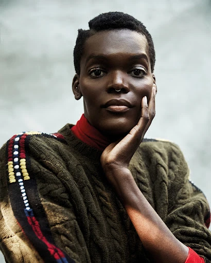 Portrait of a dark-skinned person with a nose ring, wearing a green sweater with decorative shoulder stripes, hand to face, looking at camera.