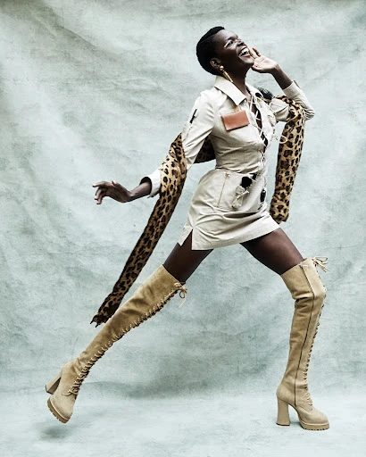 Glamorous dark-skinned woman poses in thigh-high lace-up boots, a safari-style dress, and a leopard stole in a studio setting.