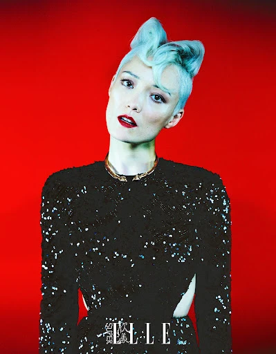 Elegant adult woman in a black sequined top with sculptural blue hair posing against a vibrant red backdrop in a fashion editorial.
