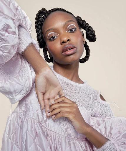 Portrait of a young Black woman with creative white eyeliner and braided pigtails wearing a puffy pink coat and light sweater against a beige background.