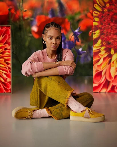 Woman with cornrow braids in a pink sweater, yellow pants, and loafers, sitting in front of a red and blue floral background.
