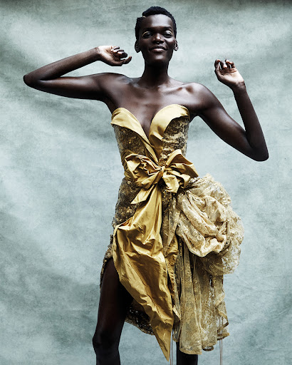 Woman in golden strapless dress with large bow and lace detail posing in a studio with arms raised, smiling in a medium shot.