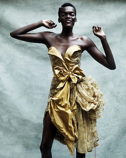 Woman in golden strapless dress with large bow and lace detail posing in a studio with arms raised, smiling in a medium shot.