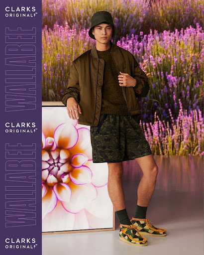 A young adult male stands in a green jacket, patterned shorts, bucket hat and Clarks shoes, leaning against a floral framed picture.