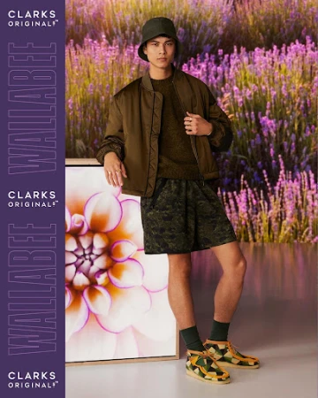 A young adult male stands in a green jacket, patterned shorts, bucket hat and Clarks shoes, leaning against a floral framed picture.