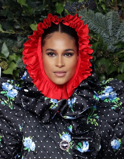 Close-up shot of a woman with fair skin wearing a polka dot floral jacket with a red ruffled hood against a greenery backdrop.